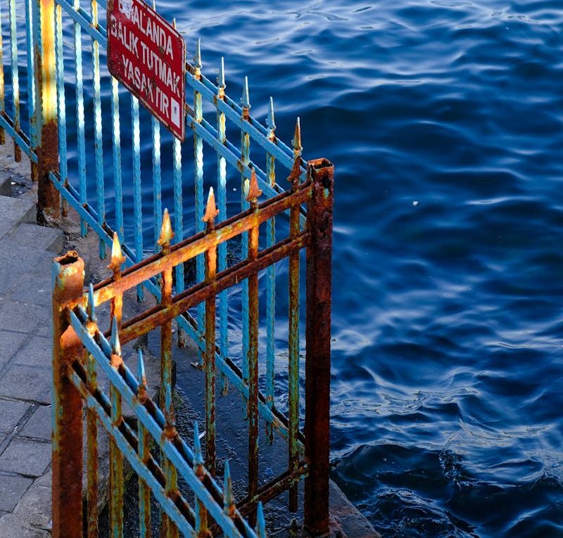 Rusty metal railings next to sea with warning sign in Turkish.